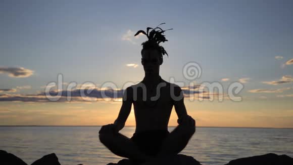 日落时分一个头上戴着美洲印第安人羽毛摩霍克配件的男子练习瑜伽的剪影视频的预览图