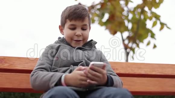 小男孩在公园里用电话他在秋天的天气里坐在长凳上背景黄树视频的预览图