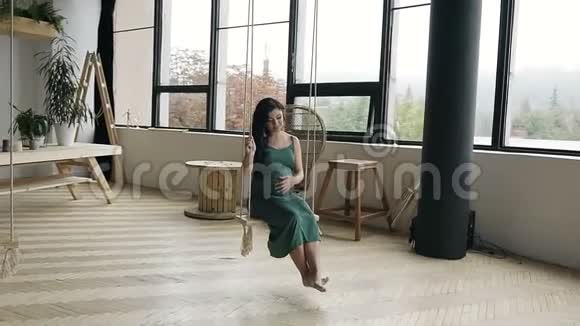 年轻的怀孕女孩在木制秋千上摇摆一个年轻的女孩穿着绿色的长裙在工作室的秋千上视频的预览图