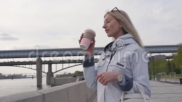 年轻漂亮的金发女郎喝着咖啡在河堤上散步慢动作视频的预览图