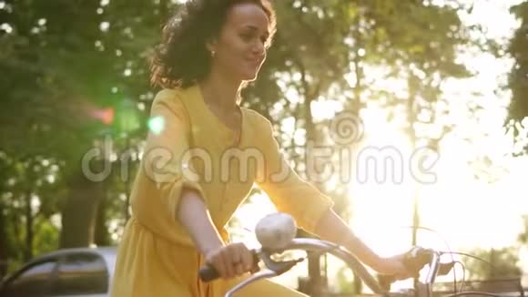 镜头耀斑微笑的快乐女人穿着黄色长裙骑着一辆城市自行车带着篮子带着鲜花在公园里视频的预览图