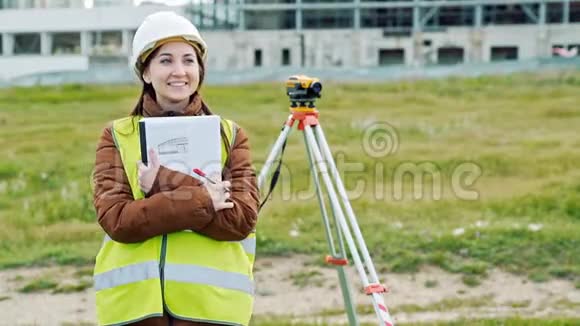 年轻的微笑女测量员穿着绿色工作服和头盔调整设备计算和书写视频的预览图