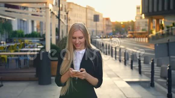 正式的女商人在街上散步优雅的金发女郎穿着西装走在街上用智能手机浏览视频的预览图