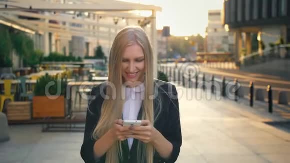 正式的女商人在街上散步优雅的金发女郎穿着西装走在街上用智能手机浏览视频的预览图