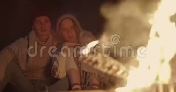 一对年轻夫妇晚上坐在森林里在篝火前的一个普通格子下拥抱谈笑风生视频的预览图