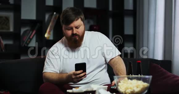胖子看电视在他的智能手机里打字他点了家常菜营养不良食物的概念视频的预览图