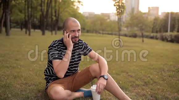 一个男人坐在绿色公园的草坪上在日落时分喝着饮料打着电话视频的预览图