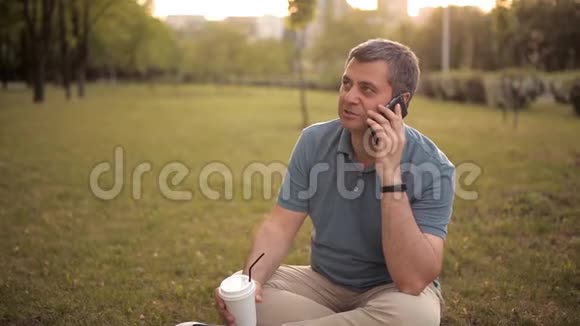 一个男人坐在绿色公园的草坪上在日落时分喝着饮料打着电话视频的预览图