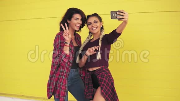 两个时髦的女孩自拍和显示和平标志站在黄色的墙美丽的女孩摆姿势和拍照视频的预览图