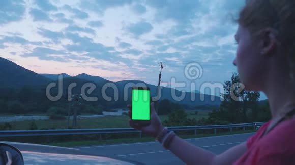 年轻的金发女郎拿着智能手机绿色屏幕在道路的背景下汽车经过视频的预览图