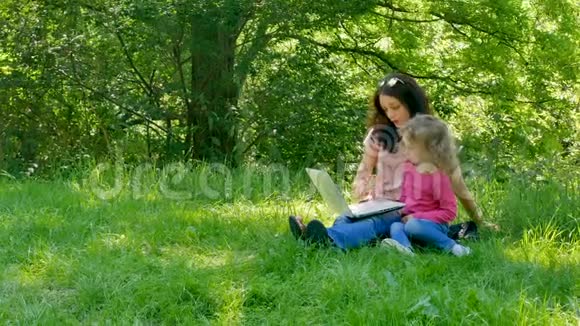 快乐家庭肖像户外在城市公园与白色笔记本电脑可爱的金发小女孩学会视频的预览图