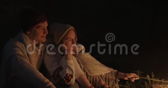 一对年轻夫妇晚上坐在森林里在篝火前的一个普通格子下拥抱谈笑风生视频的预览图