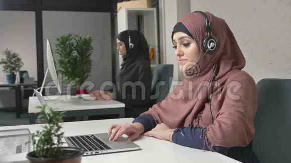 一个穿着粉红色头巾的年轻漂亮女孩正在耳机上说话在呼叫中心接听电话阿拉伯妇女参与视频的预览图