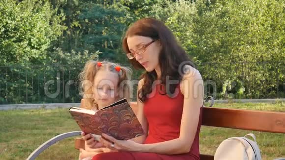 小可爱的女儿有两个小辫子和她的母亲戴着眼镜坐在公园的长凳上看书视频的预览图