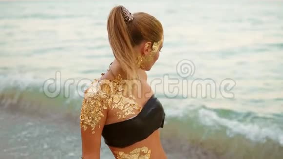 美丽的女人带着专业的金色妆容站在沙滩上的水中看着镜头视频的预览图