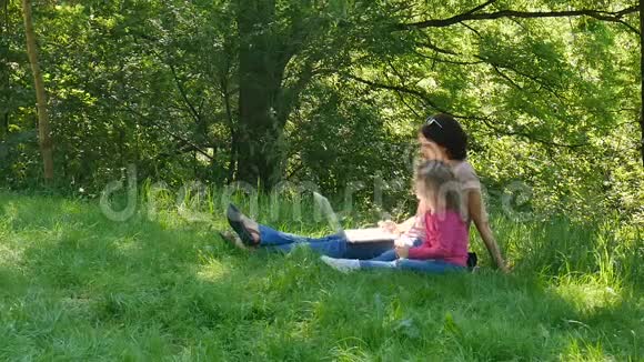 年轻的黑发母亲和金发卷发的可爱女儿正在使用白色笔记本电脑坐在绿草上视频的预览图