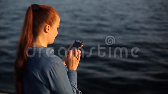 年轻漂亮的白种人女人红头发坐在海边的一块大石头上手里拿着电话视频的预览图