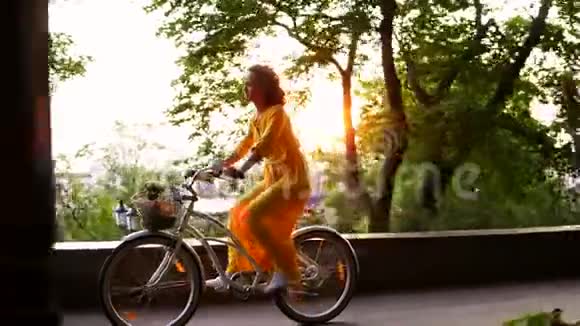 布鲁内特带着微笑的女人穿着一件黄色长裙正享受着骑着一辆自行车的时光车里放着篮子和鲜花视频的预览图
