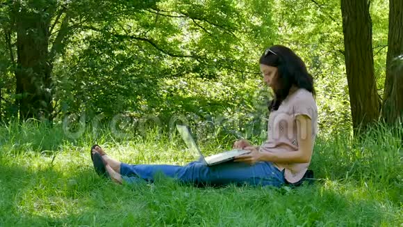 户外照片年轻的布鲁内特女士与白色笔记本电脑坐在公园的绿草上与欢乐一起工作视频的预览图