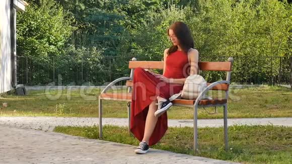 漂亮的女学生戴着眼镜穿着红长衣坐在公园的长凳上在公园里写东西视频的预览图