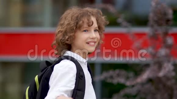 一个蓝眼睛卷曲的小学生的特写肖像男孩挥手笑了快乐的小学生回到学校视频的预览图