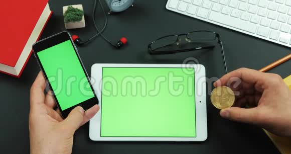 男人拿着绿色屏幕的智能手机桌子上有绿色屏幕右手拿着比特币视频的预览图