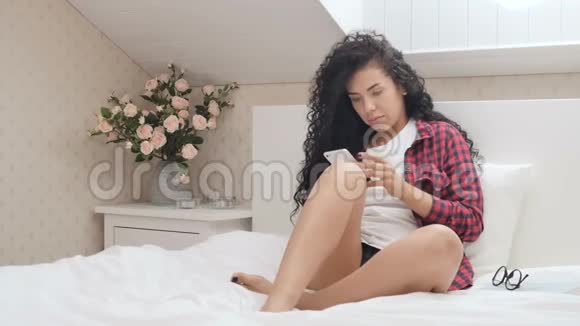 漂亮的卷发女人穿着衬衫在床上用电话视频的预览图