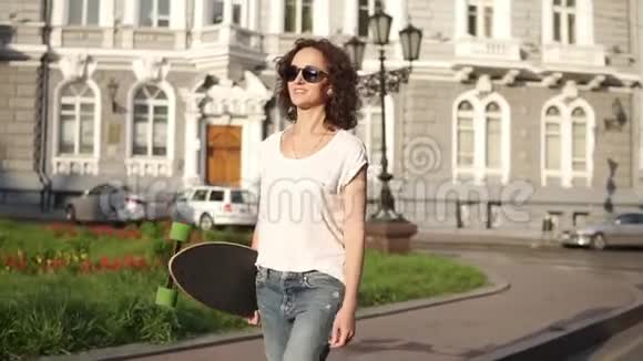 早晨黎明时分戴着墨镜的年轻美女举着长板在旧城街道散步视频的预览图