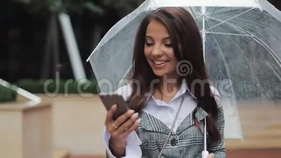 快乐的年轻商务女性使用智能手机在雨天走在街上微笑着拿着雨伞视频的预览图