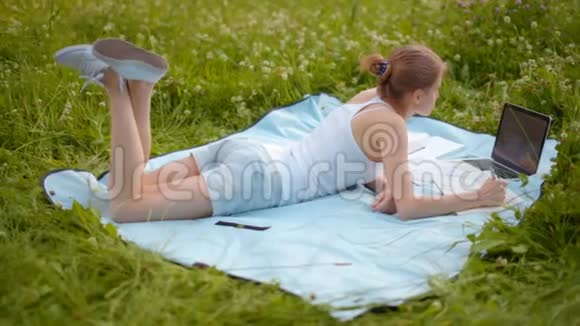 漂亮的学生女孩正在公园学习夏天在草地上用笔记本电脑和笔记本视频的预览图