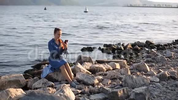 年轻漂亮的白种人女人红头发坐在海边的一块大石头上手里拿着电话视频的预览图