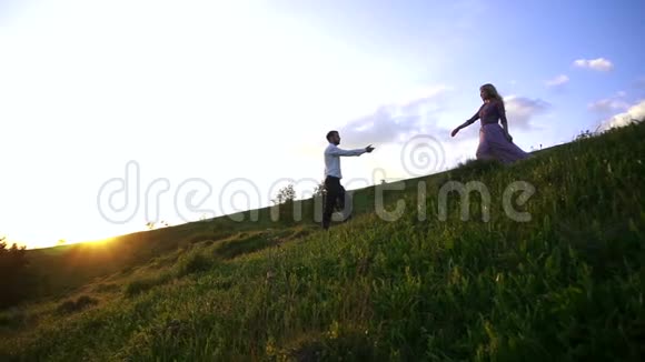 年轻迷人的金发女人穿着紫色的长裙子正在向她的爱人伸出手同时沿着草地走去他们视频的预览图