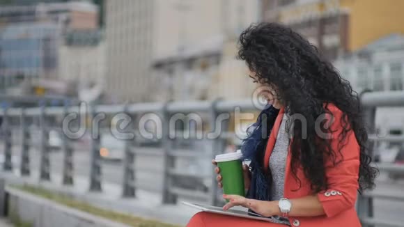 时尚的卷发女士喝咖啡在城市道路背景下工作视频的预览图