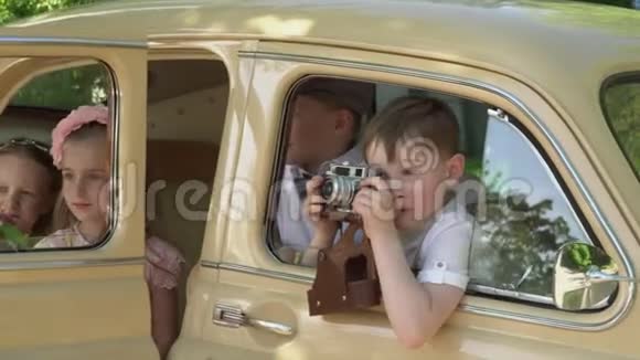 这个男孩玩一个复古的相机倾斜在米色复古车的窗户外小孩子的大公司视频的预览图