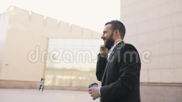 在现代办公大楼附近一个长着胡须的年轻商人拿着一杯咖啡在智能手机上交谈视频的预览图