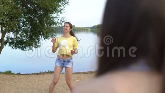 年轻女子和她的朋友在湖边的沙滩上打排球穿短T恤的漂亮女孩在玩视频的预览图