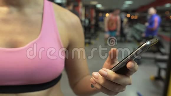 年轻女性在健身房滚动智能手机屏幕设置培训应用程序视频的预览图