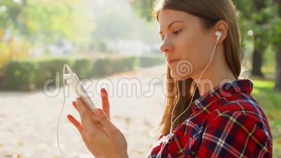 微笑的女人享受大自然坐在绿色公园的长椅上用她的手机听音乐视频的预览图