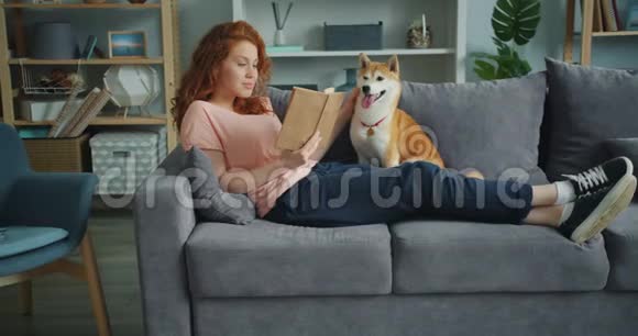 漂亮的年轻女人在家里看书爱抚坐在沙发上的狗视频的预览图
