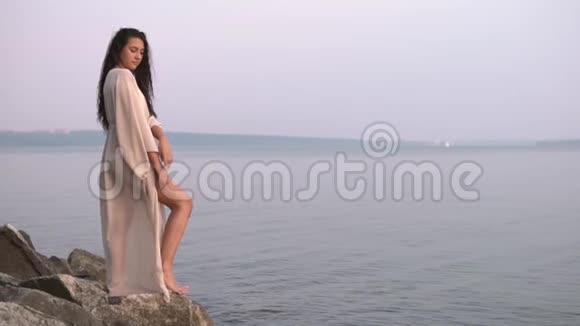 一个穿着长裙的女孩站在靠海的石头上慢动作视频的预览图