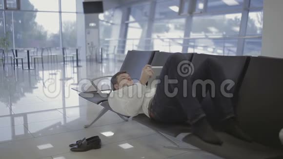 商人躺在机场候机大厅的座位上用平板电脑视频的预览图