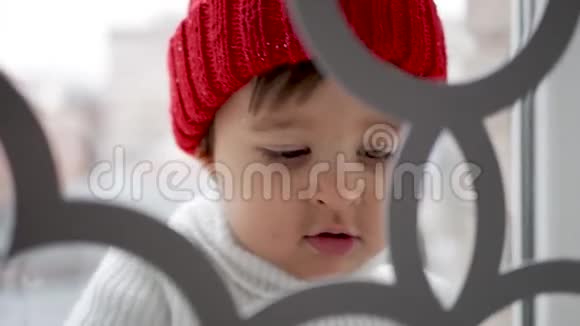 一个穿着白色针织毛衣和一顶暖红色帽子的孩子站在窗边视频的预览图