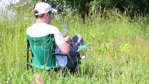 一个人坐在一片灯火通明的草地上的野餐椅上休息喝瓶子里的水从夏天开始草本植物和花朵摇摆视频的预览图