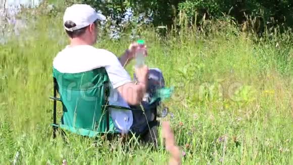 一个人坐在一片灯火通明的草地上的野餐椅上休息喝瓶子里的水从夏天开始草本植物和花朵摇摆视频的预览图