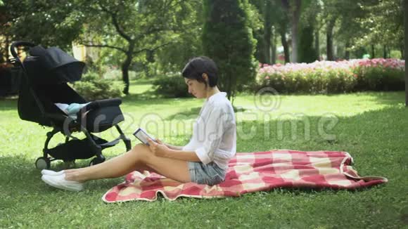 带着婴儿车的漂亮女人在公园的草地上看书视频的预览图