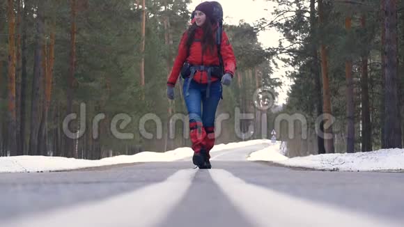 年轻女子背着背包在美丽的冬林中徒步走在柏油路上视频的预览图