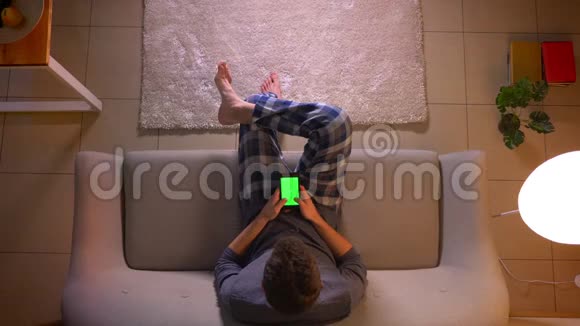 在舒适的家中年轻人坐在沙发上用绿色屏幕的手机进行特写镜头视频的预览图