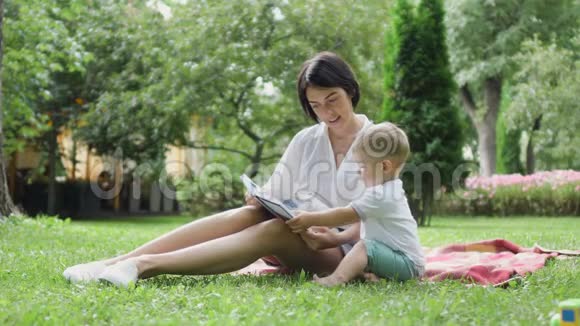 可爱的男孩和妈妈看着书坐在公园的格子呢视频的预览图