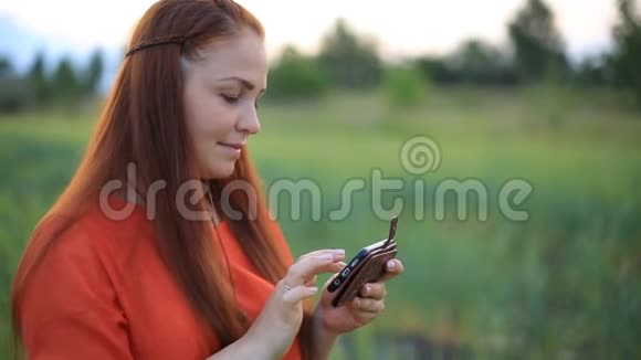 年轻漂亮的女人穿着红色的裙子留着红色的头发在你的手机上看你的照片站在绿色的麦田里视频的预览图