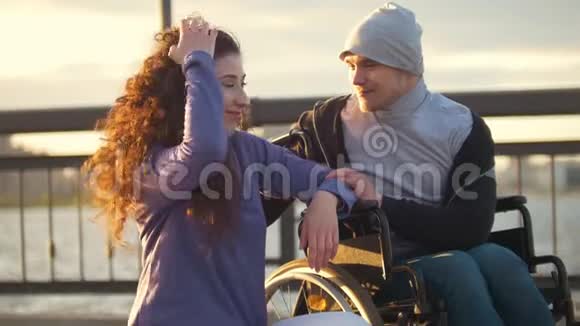 快乐的一对夫妇坐在轮椅上的残疾男子和迷人的年轻女子在夕阳下交谈视频的预览图
